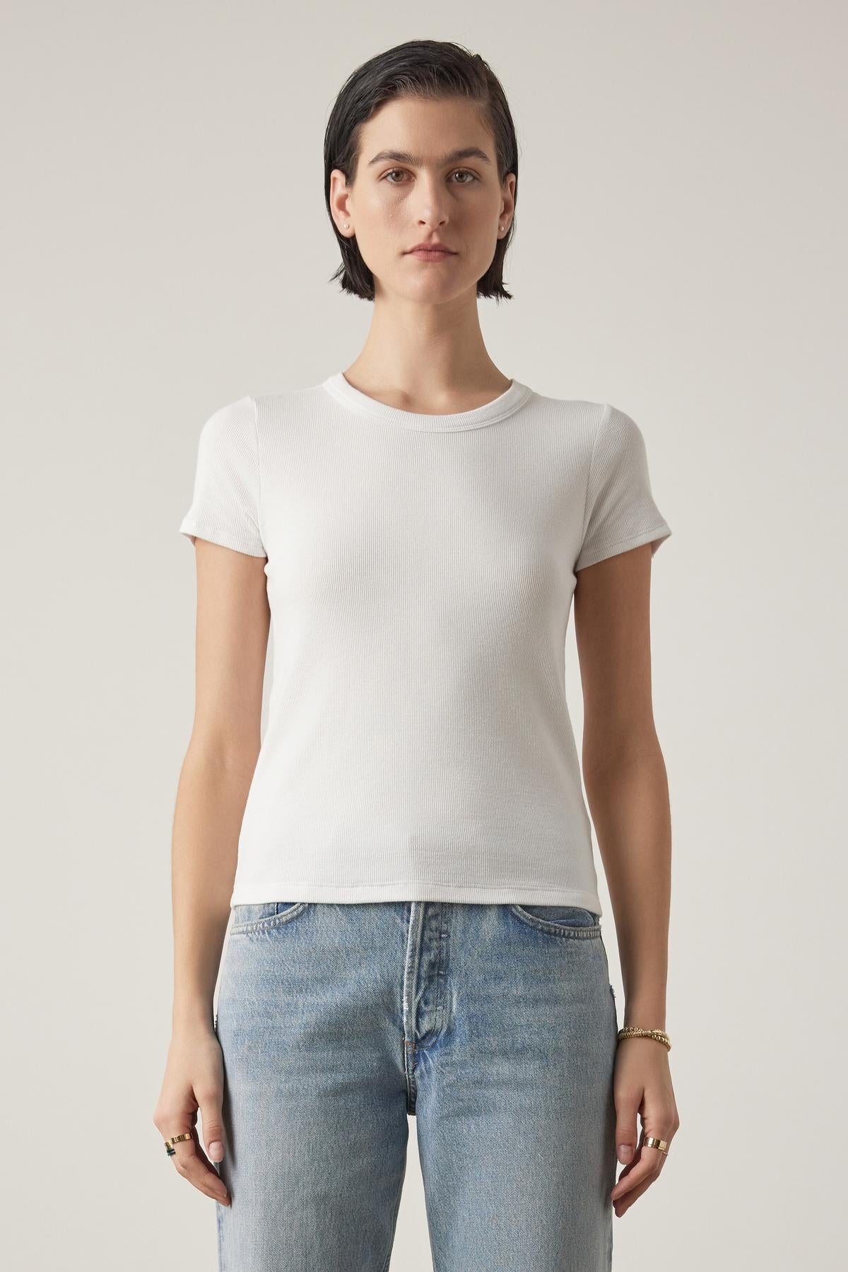 A woman with short dark hair wearing a BEDFORD TEE by Velvet by Jenny Graham in white with a crew neckline and blue jeans stands against a neutral background, facing the camera with a neutral expression.-36753616765121