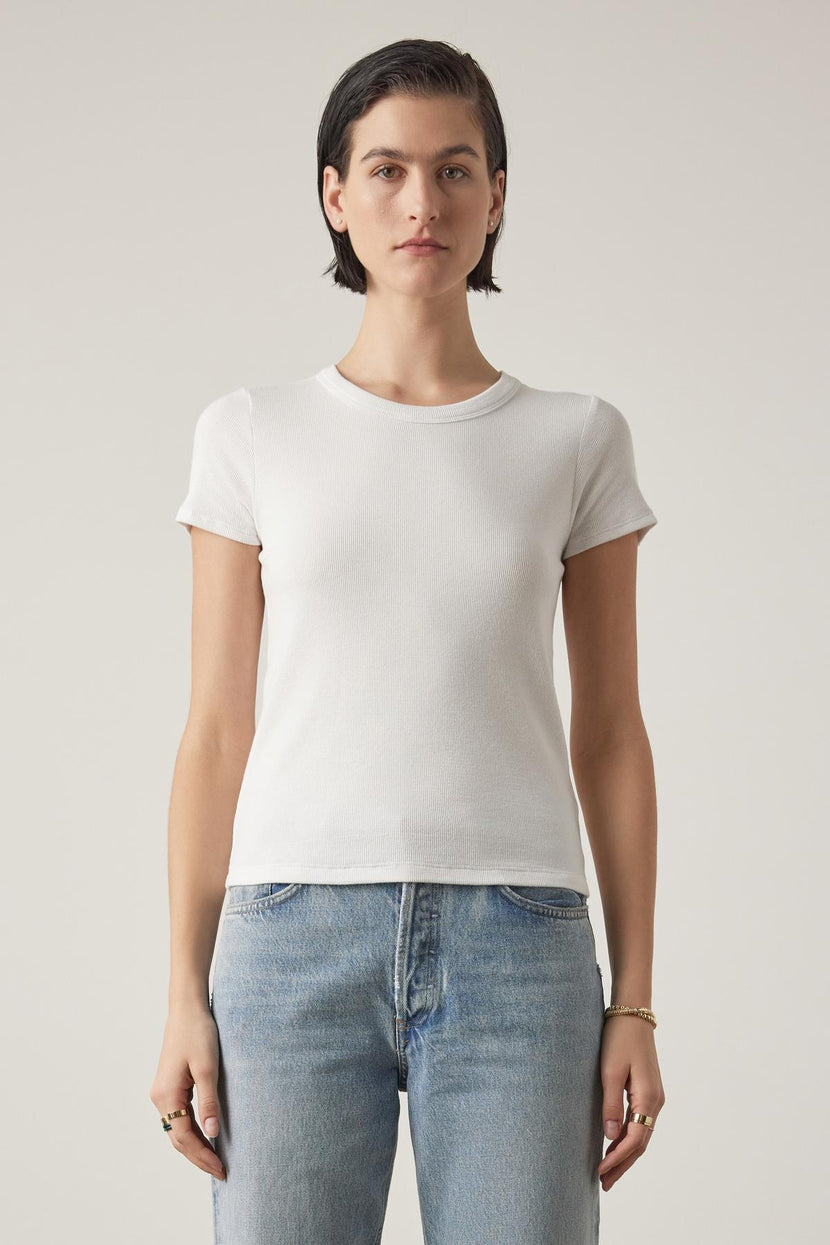 A woman with short dark hair wearing a BEDFORD TEE by Velvet by Jenny Graham in white with a crew neckline and blue jeans stands against a neutral background, facing the camera with a neutral expression.
