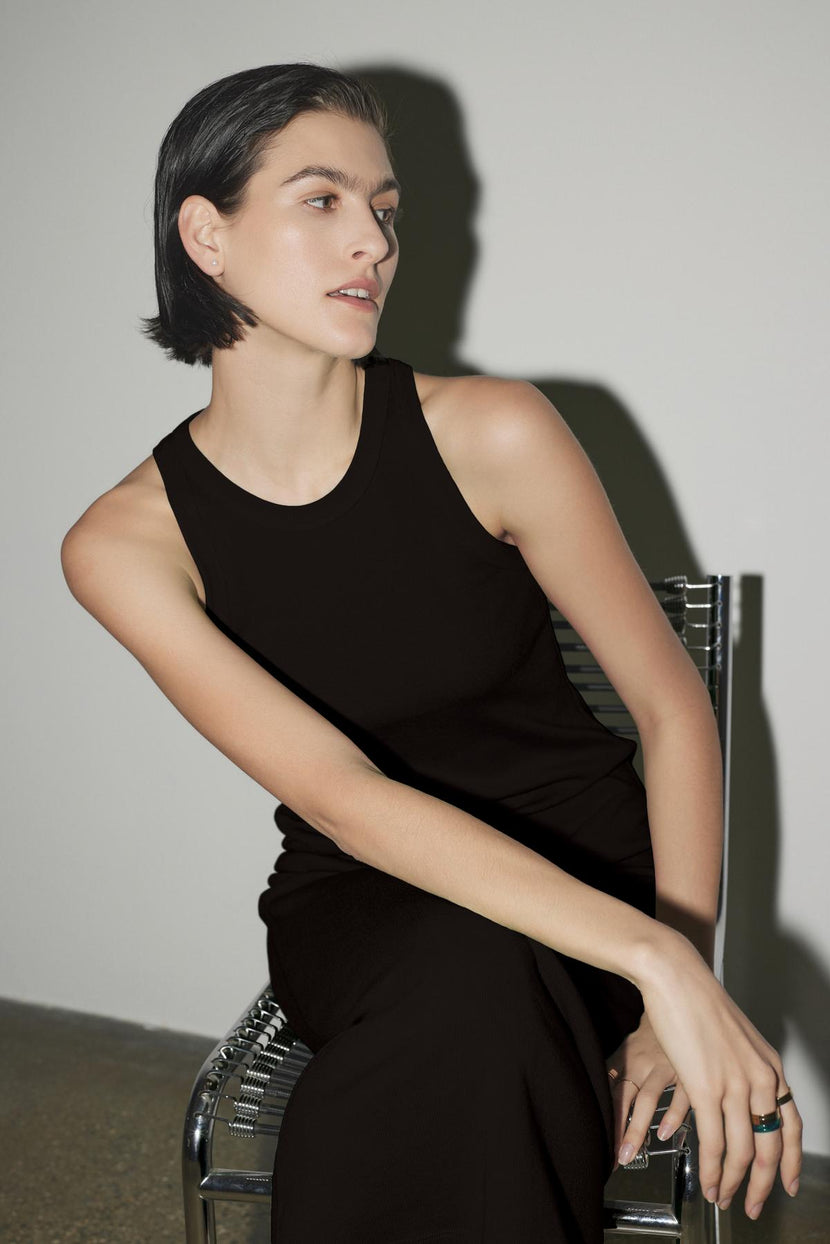 A woman with short dark hair sits on a metal chair, wearing the Velvet by Jenny Graham Griffith Dress, a black sleeveless top with a crew neckline, looking to the side in a neutral indoor setting.