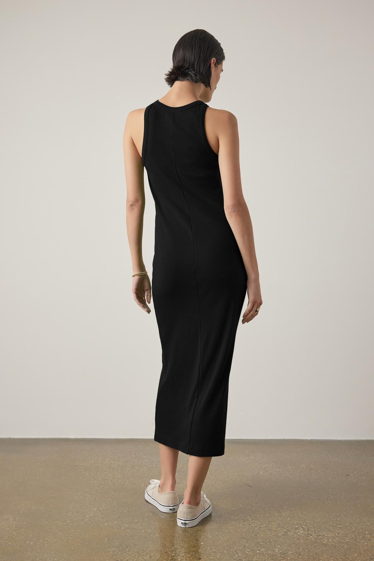 A woman in a sleek black sleeveless GRIFFITH DRESS from Velvet by Jenny Graham with a ribbed texture, seen from the back, paired with white sneakers, standing in a simple room with a concrete floor.-36891634368705