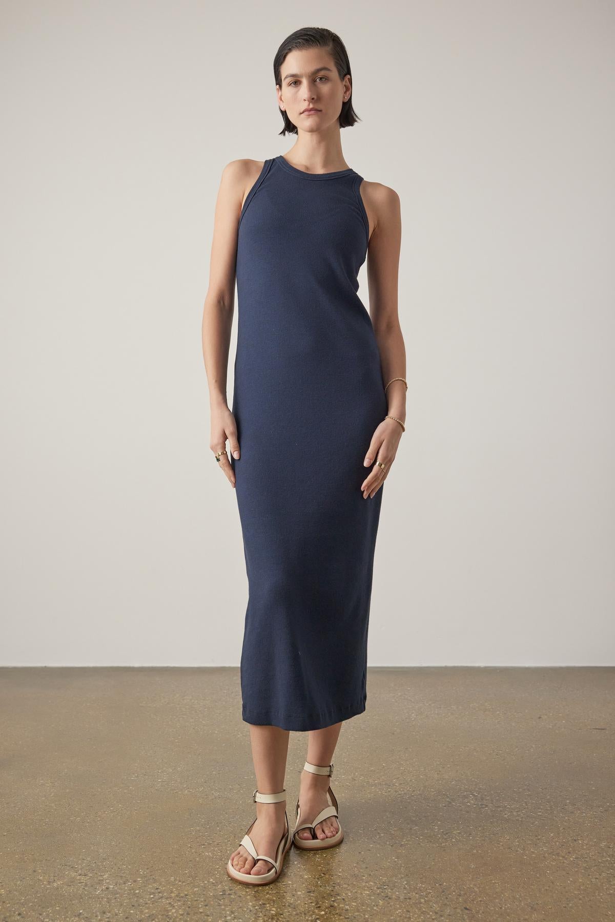 A woman in a Griffith dress by Velvet by Jenny Graham, a navy blue sleeveless midi dress with a crew neckline, stands in a neutral studio setting, paired with beige sandals.-36863292637377