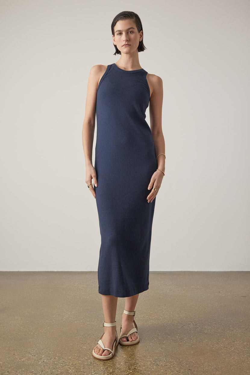 A woman in a Griffith dress by Velvet by Jenny Graham, a navy blue sleeveless midi dress with a crew neckline, stands in a neutral studio setting, paired with beige sandals.