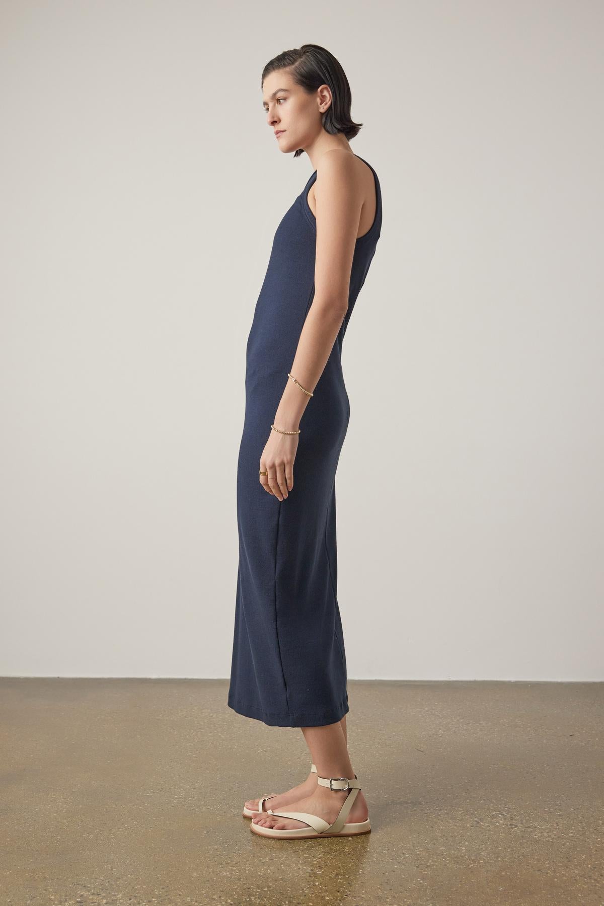   Woman in a navy blue sleeveless Griffith Dress by Velvet by Jenny Graham, with a crew neckline, standing sideways, paired with beige sandals against a neutral background. 
