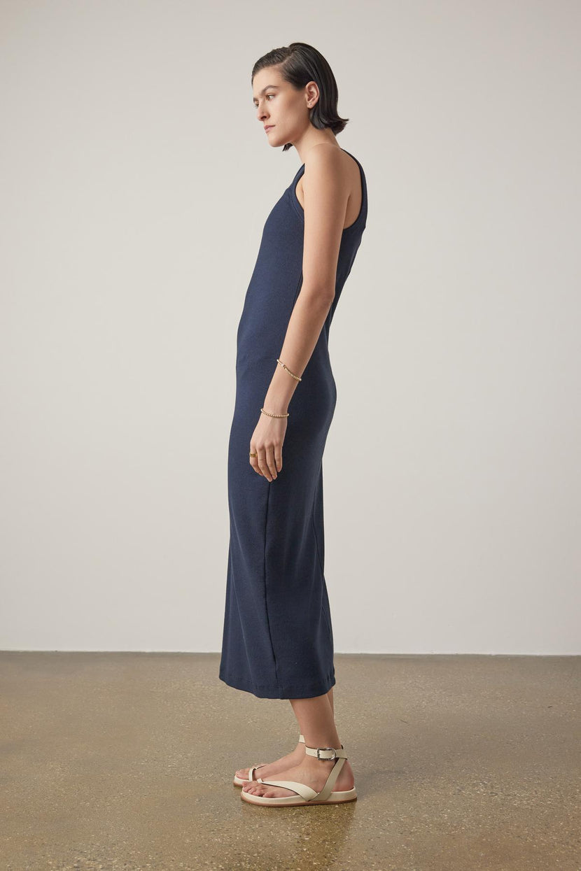 Woman in a navy blue sleeveless Griffith Dress by Velvet by Jenny Graham, with a crew neckline, standing sideways, paired with beige sandals against a neutral background.