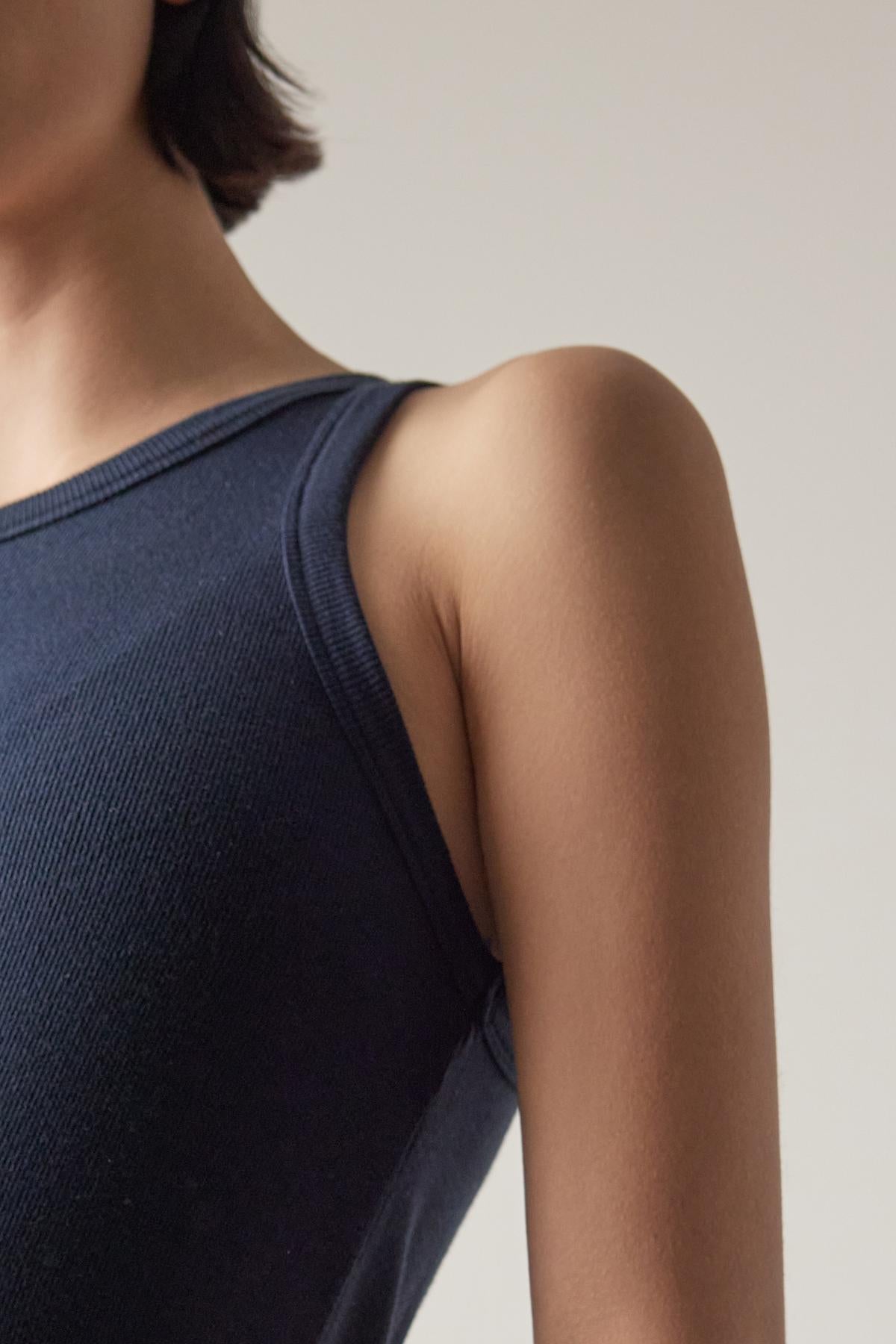 Close-up of a person wearing a dark blue tank top with a crew neckline by Velvet by Jenny Graham, focusing on the shoulder and upper arm, with a neutral beige background.-36863292801217