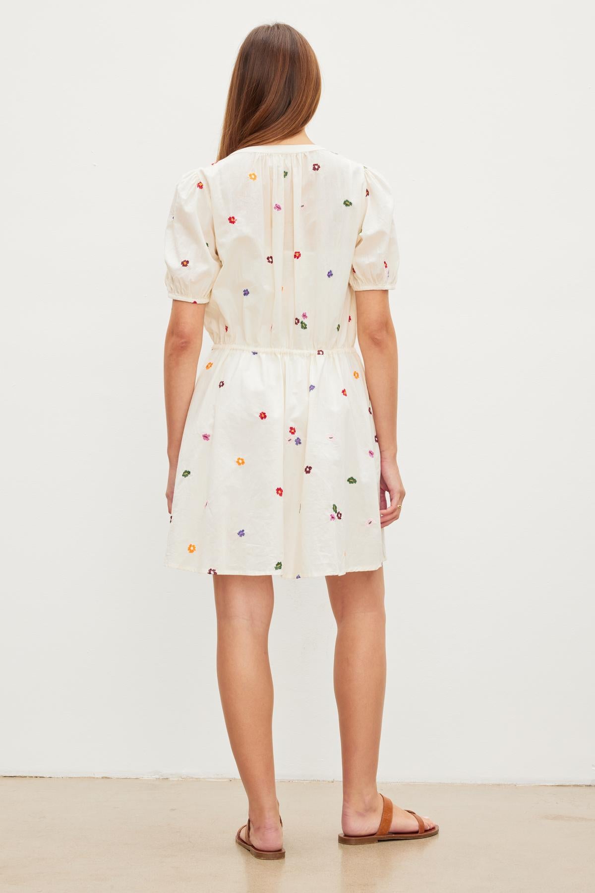 Woman standing with her back to the camera, wearing a short white CLEO EMBROIDERED BOHO DRESS by Velvet by Graham & Spencer with multicolored floral embroidery and a drawstring waist, against a plain white background.-35955682279617