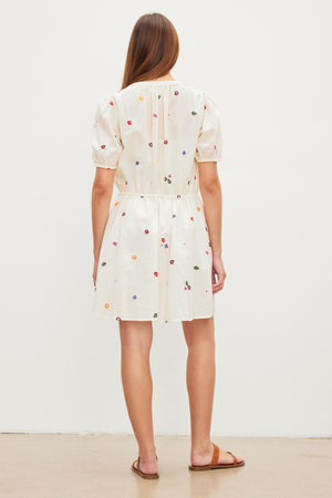 Woman standing with her back to the camera, wearing a short white CLEO EMBROIDERED BOHO DRESS by Velvet by Graham & Spencer with multicolored floral embroidery and a drawstring waist, against a plain white background.