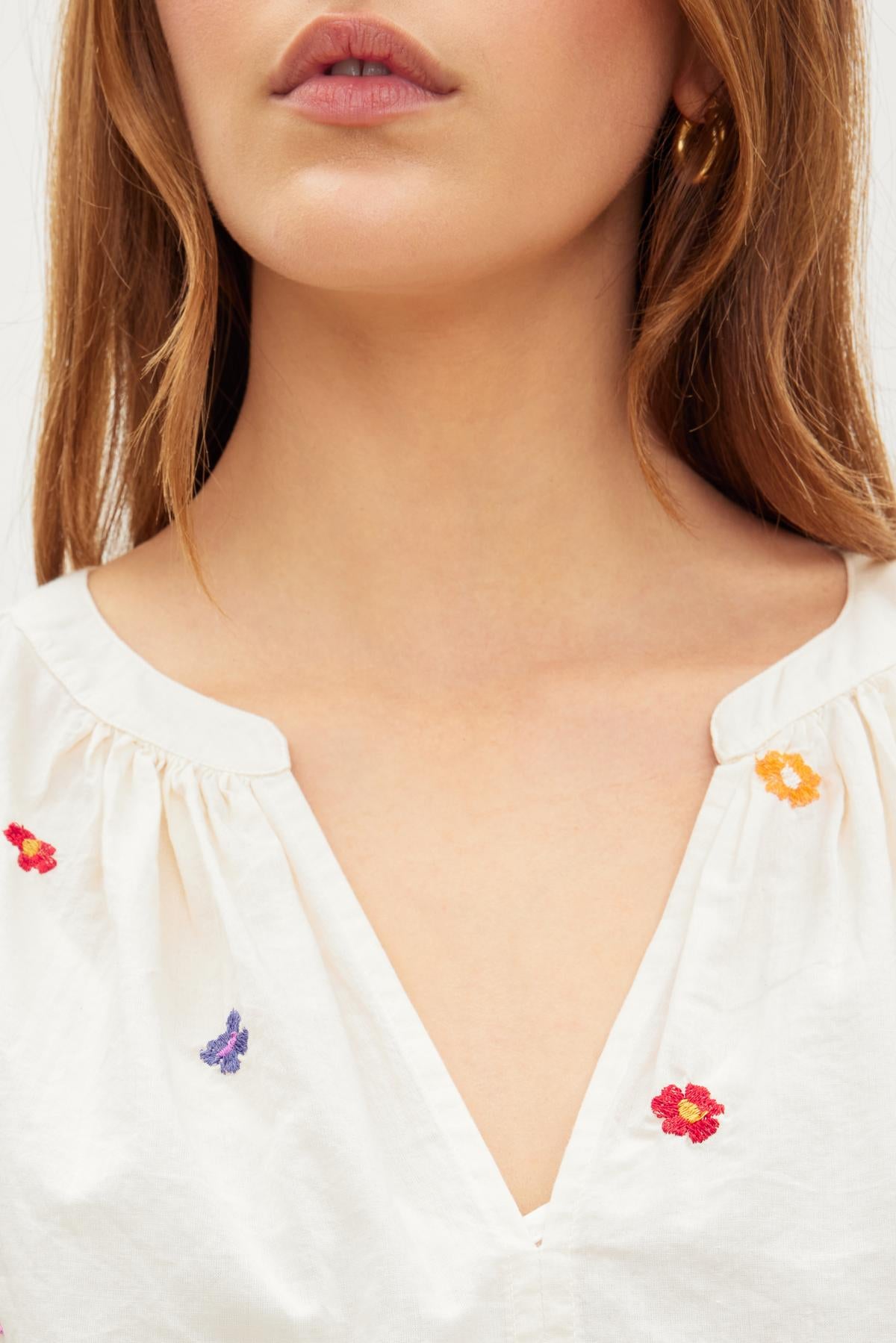   Close-up view of a woman wearing a white CLEO EMBROIDERED BOHO DRESS with a v-neckline, adorned with small embroidered flowers in orange, red, and purple by Velvet by Graham & Spencer. 