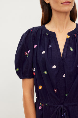 A woman in a navy Velvet by Graham & Spencer dress with colorful floral embroidery on the sleeves, chest, and a v-neckline stands against a neutral background. Only her torso and one shoulder are visible.