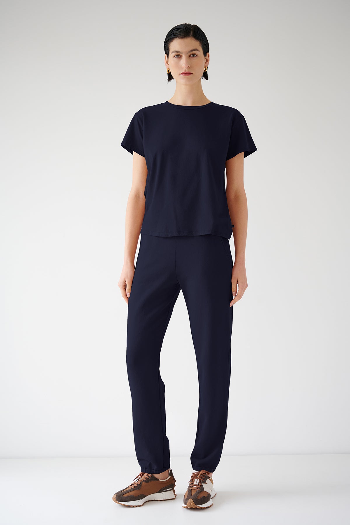 A woman in a navy blue organic cotton t-shirt and ZUMA SWEATPANT stands against a white background, wearing brown and beige sneakers by Velvet by Jenny Graham.-36891041366209