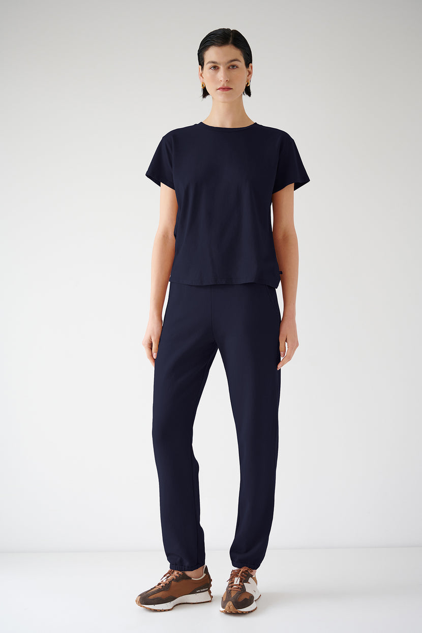 A woman in a navy blue organic cotton t-shirt and ZUMA SWEATPANT stands against a white background, wearing brown and beige sneakers by Velvet by Jenny Graham.