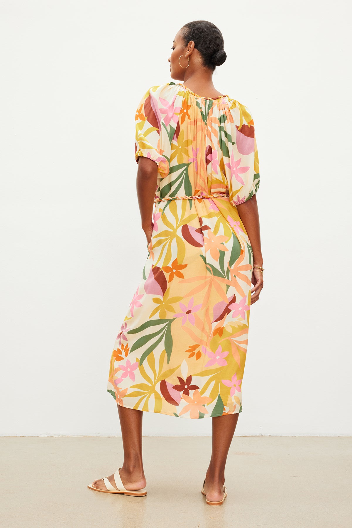 A woman stands with her back to the camera, wearing a Velvet by Graham & Spencer CAROL PRINTED BOHO DRESS with a detachable twist braid belt and tan sandals against a plain background.-35955494355137