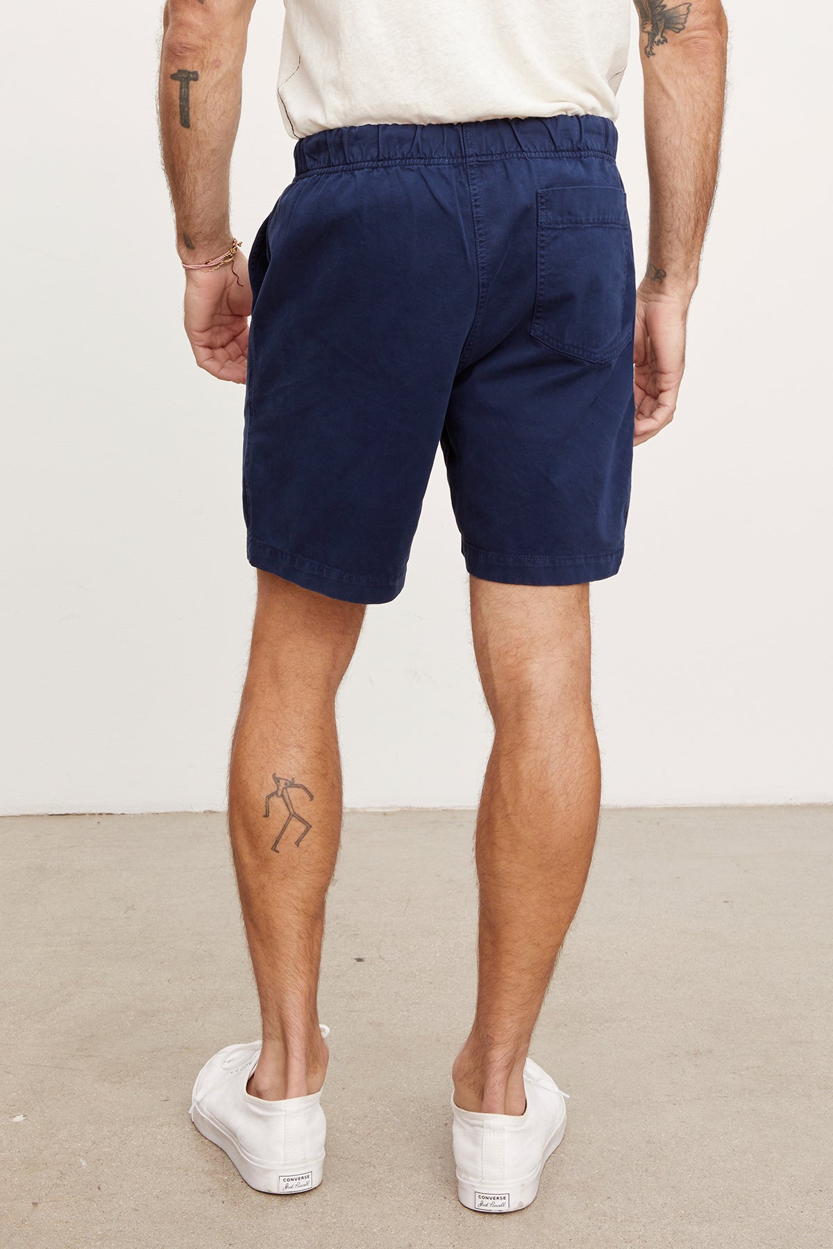   A man viewed from behind wearing navy blue cotton twill Velvet by Graham & Spencer Fielder shorts and white sneakers, standing with hands in pockets; visible tattoos on his legs. 