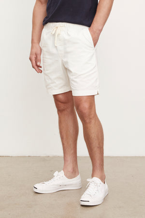 Man wearing a navy blue t-shirt and FIELDER shorts by Velvet by Graham & Spencer with white sneakers, standing in a room with a plain background. Only his torso and legs are visible.