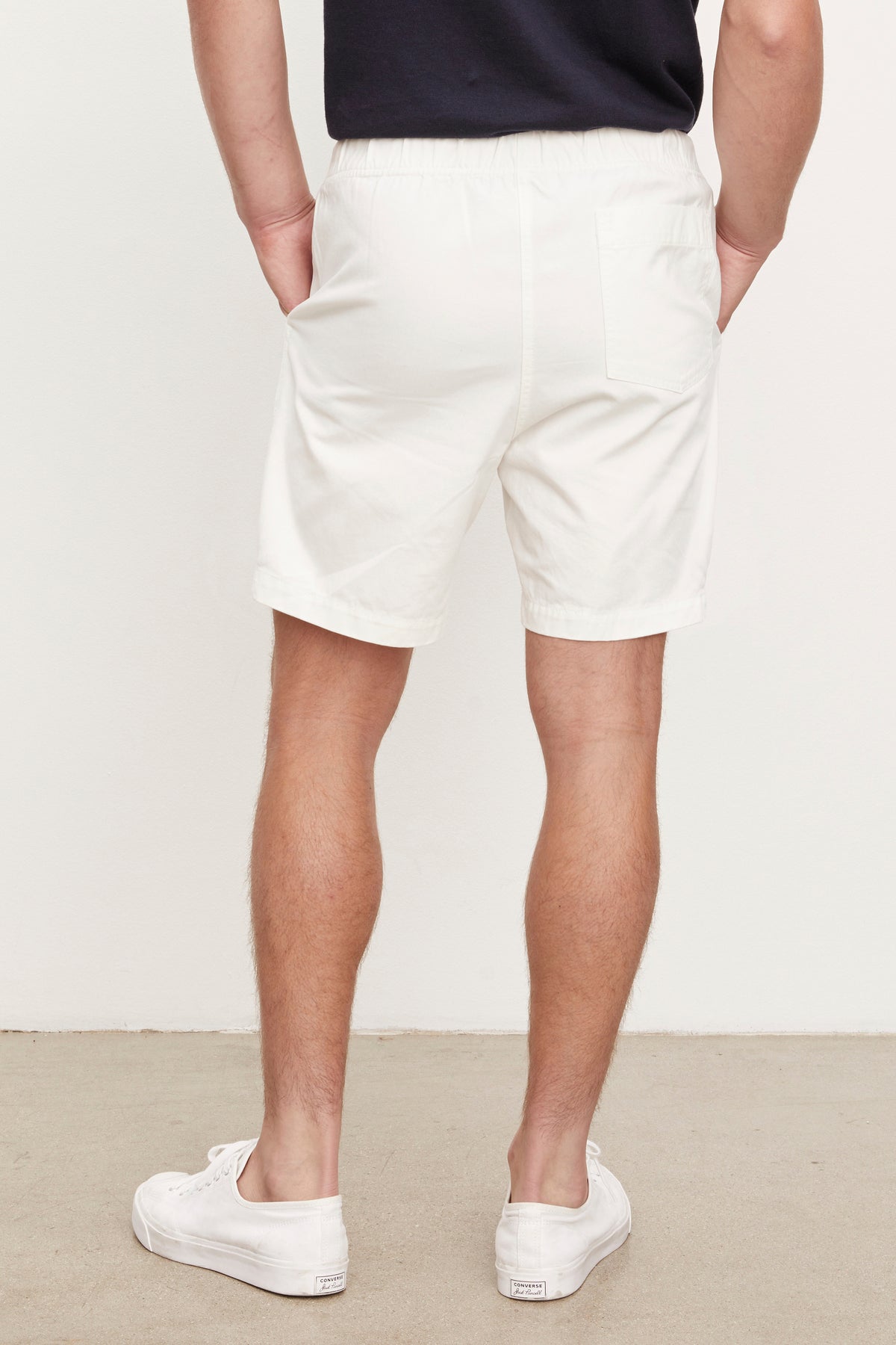 A man viewed from behind wearing white Fielder shorts and white sneakers, standing against a plain wall.-36753575903425
