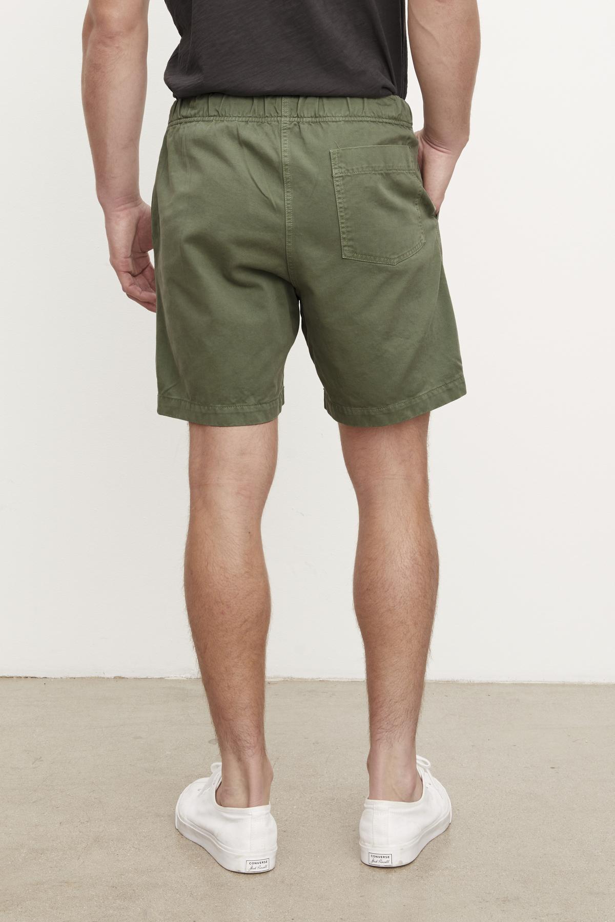   A man wearing Velvet by Graham & Spencer FIELDER SHORT in olive green cotton twill shorts and white sneakers standing with his back to the camera, indoors against a white wall. 