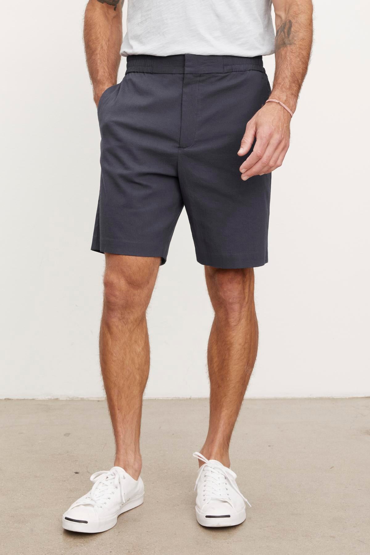   A frontal view of a man from the waist down, wearing Velvet by Graham & Spencer's DAMIAN SHORTS with slash pockets and white sneakers. He has his left hand partially in his pocket. 