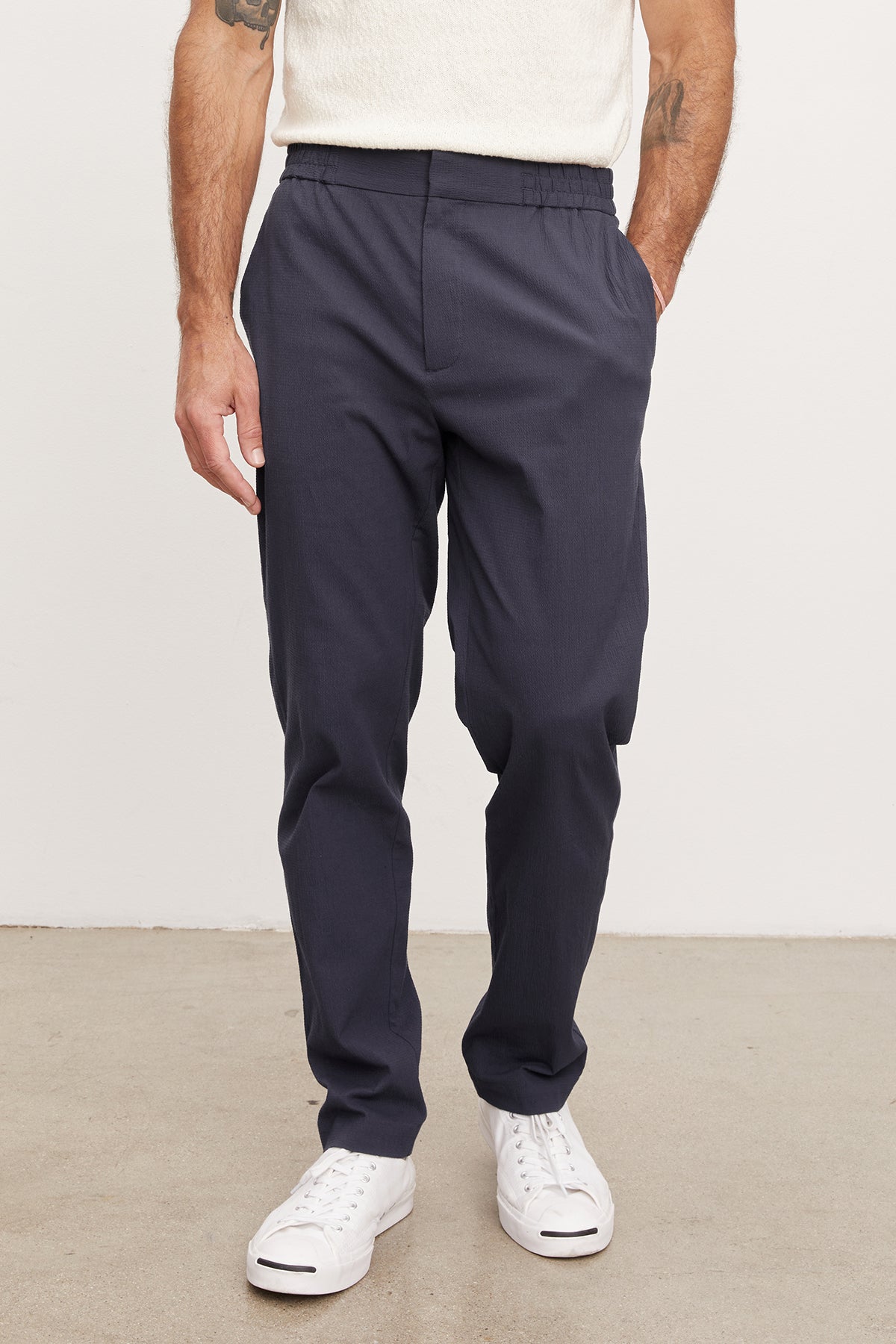 A man wearing navy blue seersucker cotton WILLEM PANTS by Velvet by Graham & Spencer and white sneakers, standing with his hands on his hips. Only the lower half of his body is visible.-36909312704705