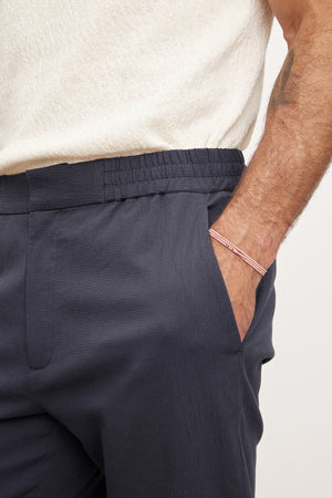 Close-up of a man's side, showing Velvet by Graham & Spencer navy blue seersucker cotton Willem pants with an elastic waistband and a single pocket, against a light beige sweater.