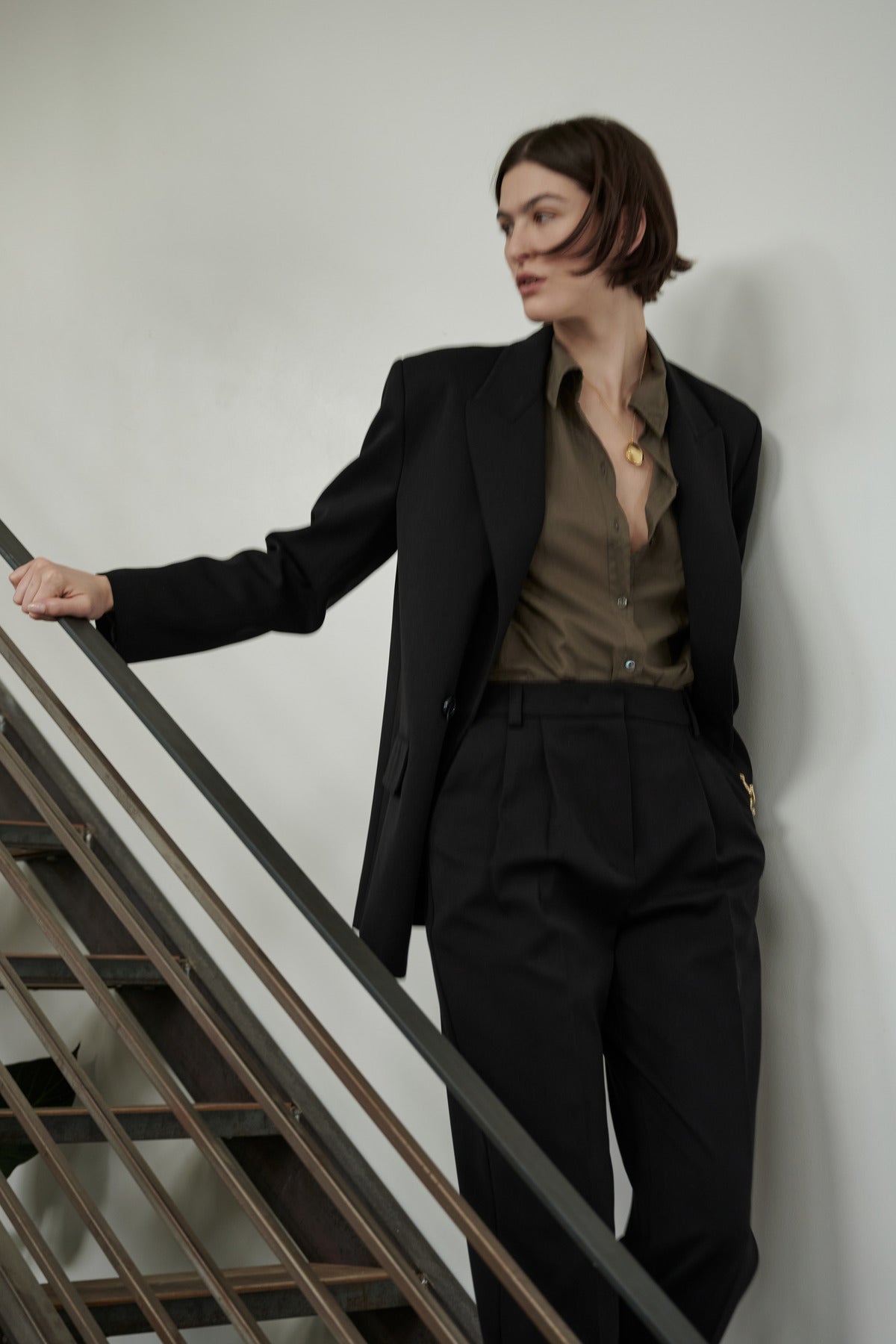 A person in a stylish Velvet by Jenny Graham FAIRFAX BLAZER with structured shoulders and olive shirt posing beside a staircase railing.-35995793948865