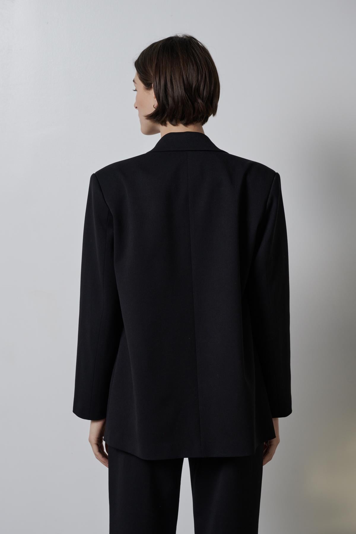 Woman standing with her back to the camera, wearing a black double-breasted Fairfax blazer with a straight-cut silhouette from Velvet by Jenny Graham.-35995793883329