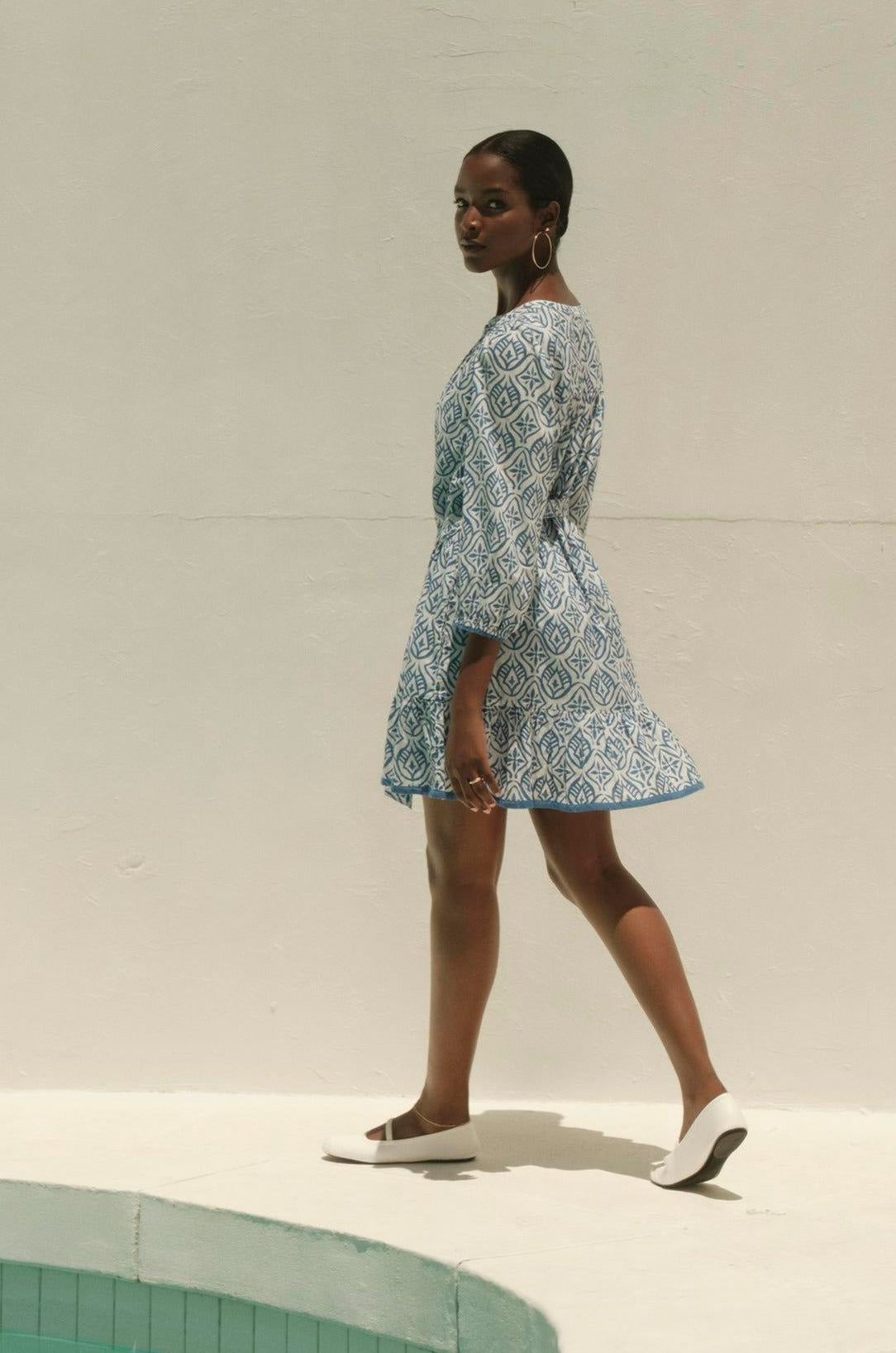 A woman in a KENLEY PRINTED BOHO DRESS by Velvet by Graham & Spencer and white shoes walks past a swimming pool, casting a long shadow on a sunlit wall.-36161169621185