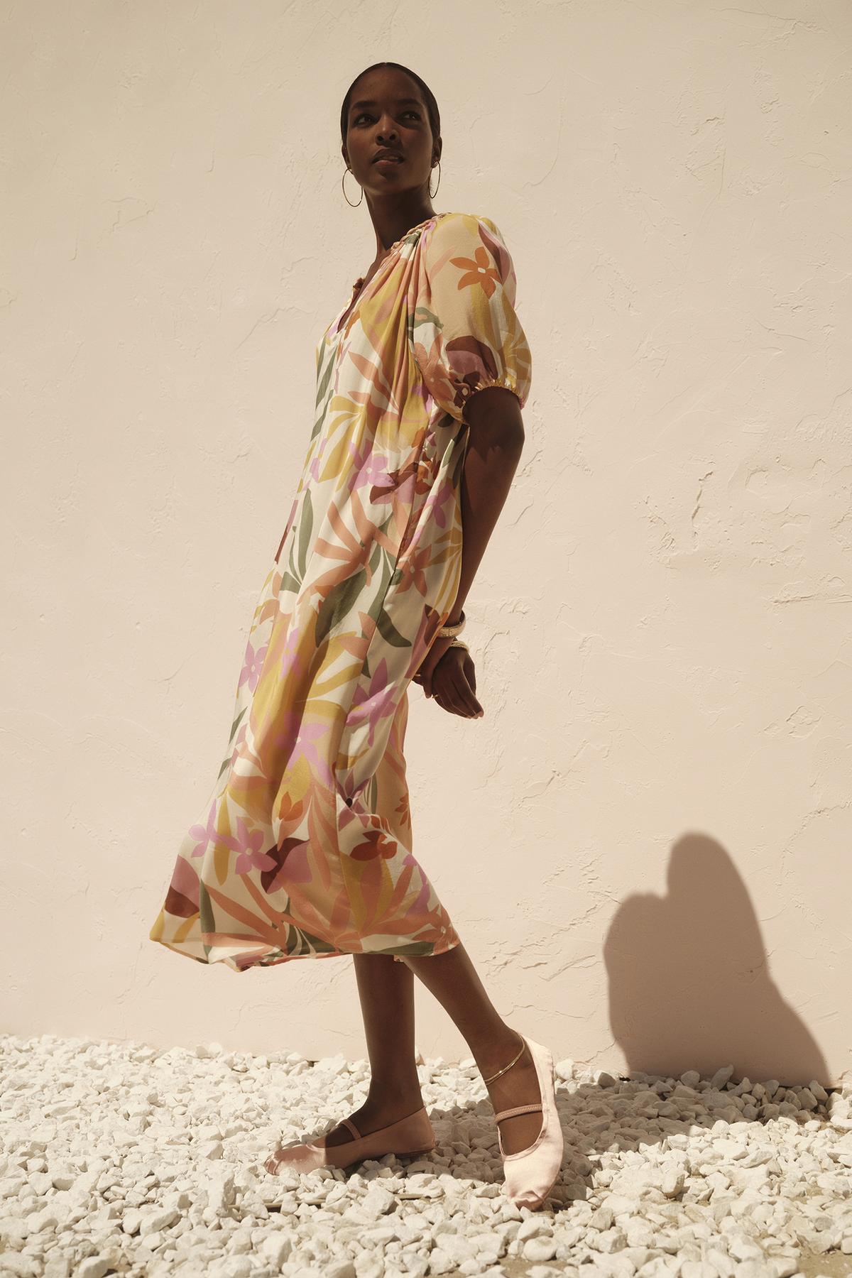 A woman in a flowing, pastel-colored CAROL PRINTED BOHO DRESS by Velvet by Graham & Spencer with a detachable twist braid belt stands against a light beige wall, casting a long shadow on a white pebbled surface.-36783538208961