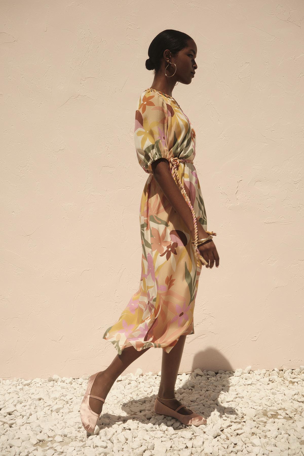 A woman in a Velvet by Graham & Spencer CAROL PRINTED BOHO DRESS with a detachable twist braid belt and pink shoes stands profiled against a pale wall, sunlight casting shadows over her and the white gravel beneath.-36783538241729