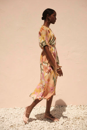A woman in a Velvet by Graham & Spencer CAROL PRINTED BOHO DRESS with a detachable twist braid belt and pink shoes stands profiled against a pale wall, sunlight casting shadows over her and the white gravel beneath.