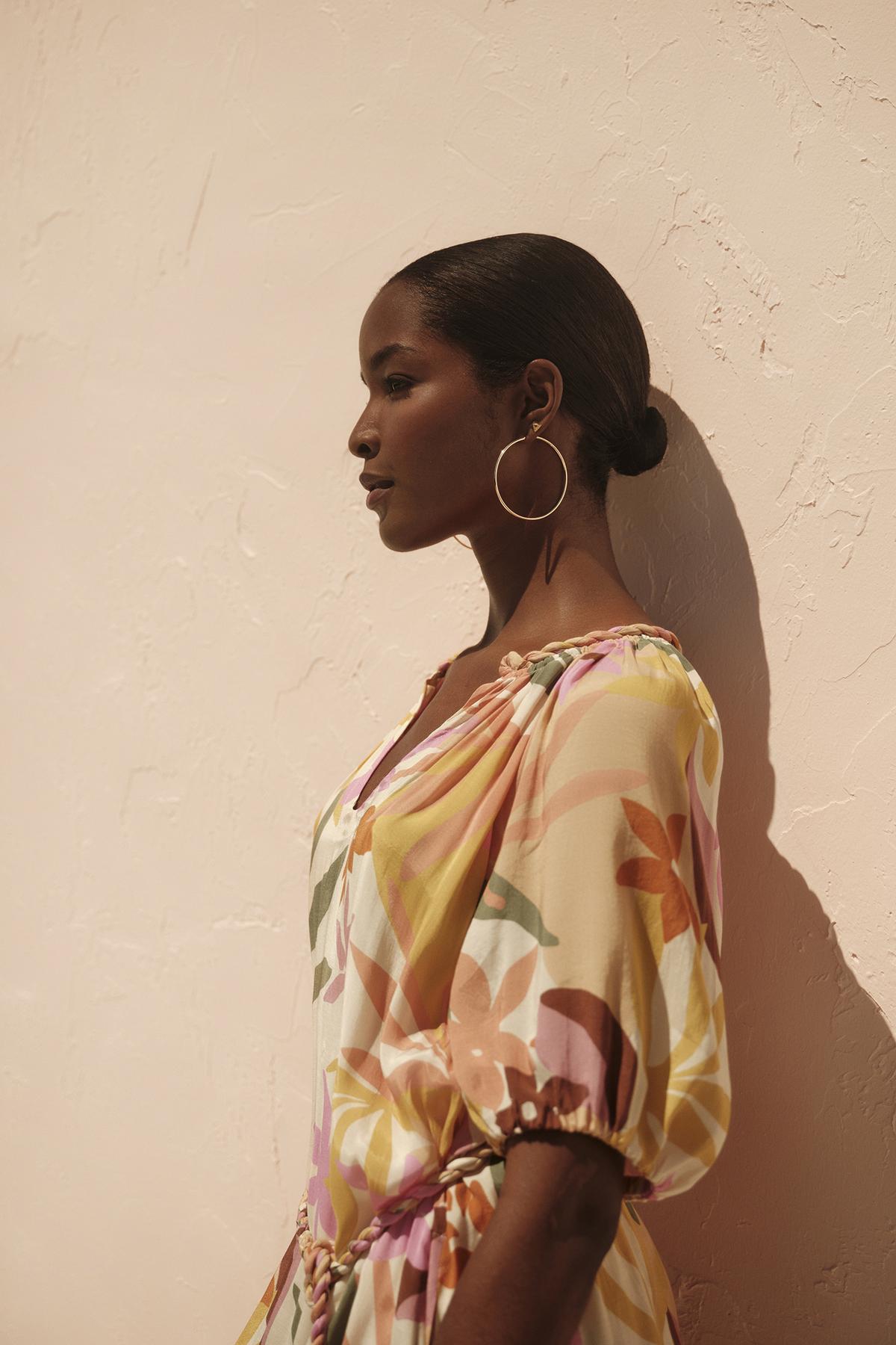   A woman in a Velvet by Graham & Spencer CAROL PRINTED BOHO DRESS stands against a textured wall, profile view, with sunlight casting shadows on her face. 