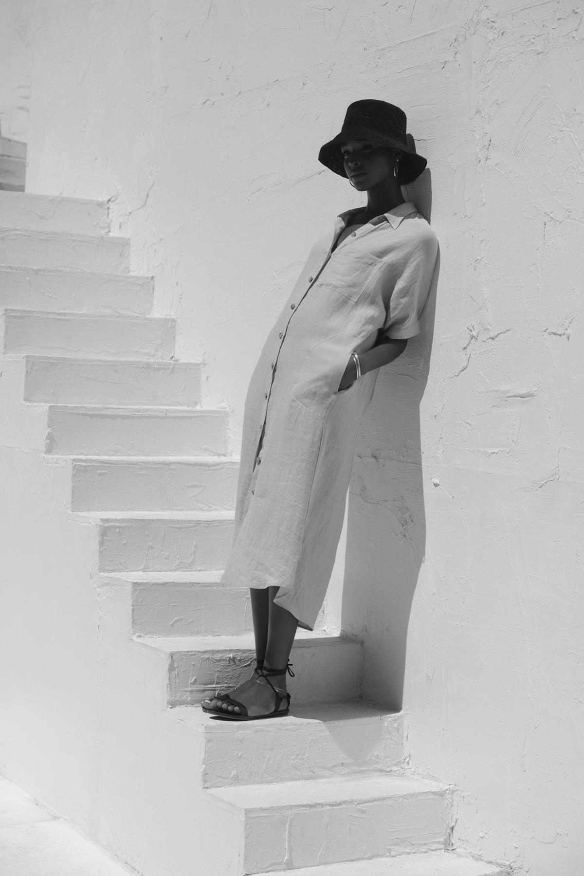   A woman in a Sandra Linen Button-Up Dress by Velvet by Graham & Spencer and wide-brimmed hat stands on white stairs against a white wall, posing in a sunny ambiance. 