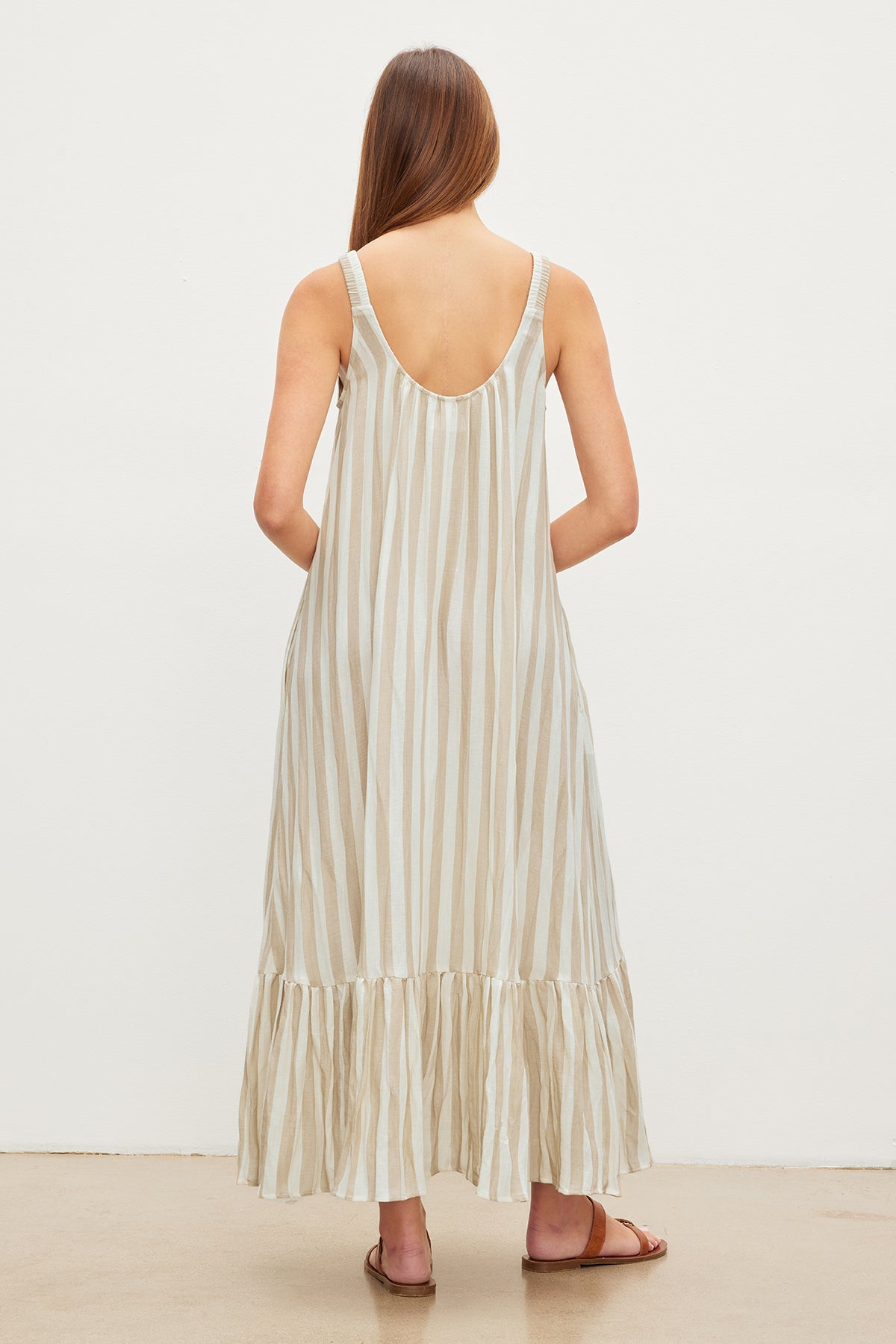 A woman stands facing away from the camera, wearing a Velvet by Graham & Spencer MERADITH STRIPED LINEN MAXI DRESS with thin straps and ruffled hem, paired with brown sandals.-35982309654721