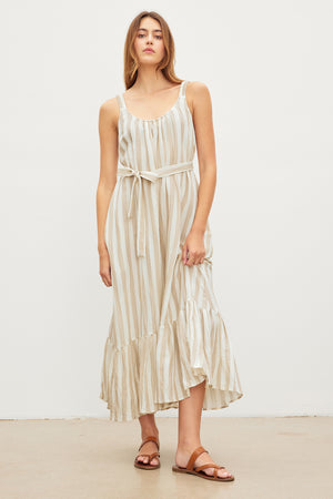 A woman stands in a studio, wearing a Velvet by Graham & Spencer MERADITH STRIPED LINEN MAXI DRESS with a belt and ruffled hem, paired with brown sandals.