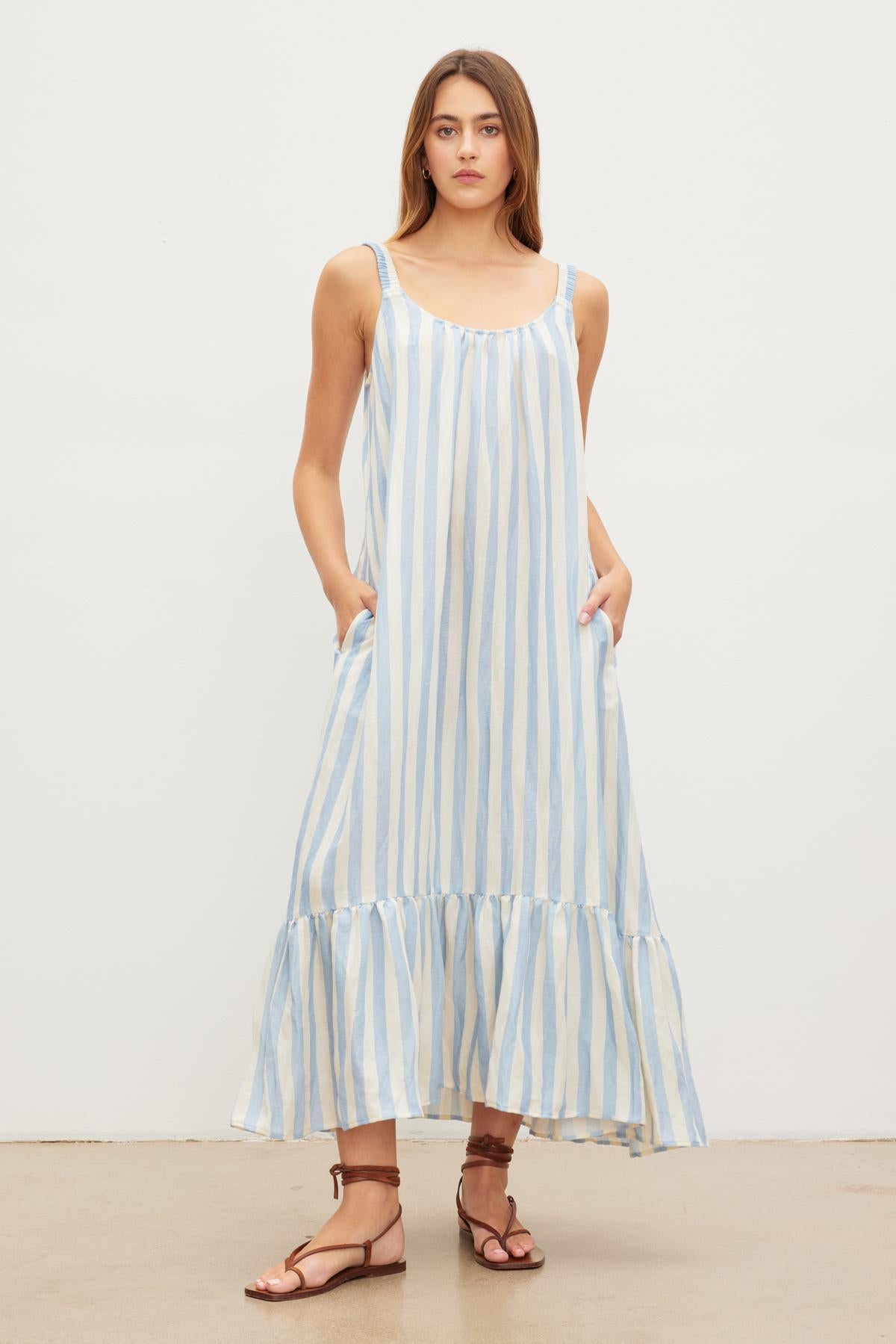 A woman stands against a plain background wearing a Velvet by Graham & Spencer MERADITH STRIPED LINEN MAXI DRESS with ruffle hem and brown sandals.-36580695441601