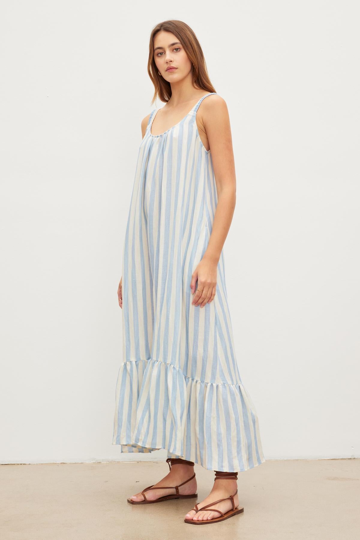 A woman stands against a plain background, wearing a Velvet by Graham & Spencer MERADITH STRIPED LINEN MAXI DRESS with thin straps and ruffled hemline, paired with brown sandals.-36580695474369