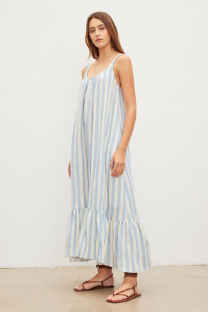 A woman stands against a plain background, wearing a Velvet by Graham & Spencer MERADITH STRIPED LINEN MAXI DRESS with thin straps and ruffled hemline, paired with brown sandals.