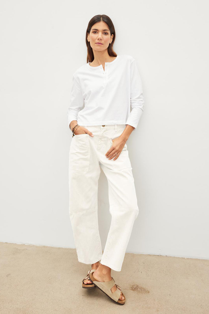 A woman stands against a plain background, wearing a white long-sleeve top, White BRYLIE SANDED TWILL UTILITY PANT with patch pockets, and brown sandals by Velvet by Graham & Spencer.