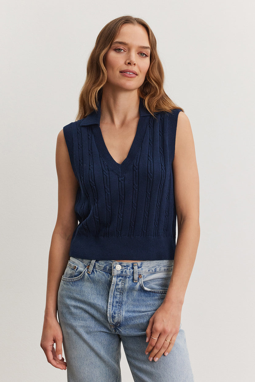 A woman wearing a navy blue WENDY SWEATER VEST and light blue jeans stands against a neutral background. Brand: Velvet by Graham & Spencer