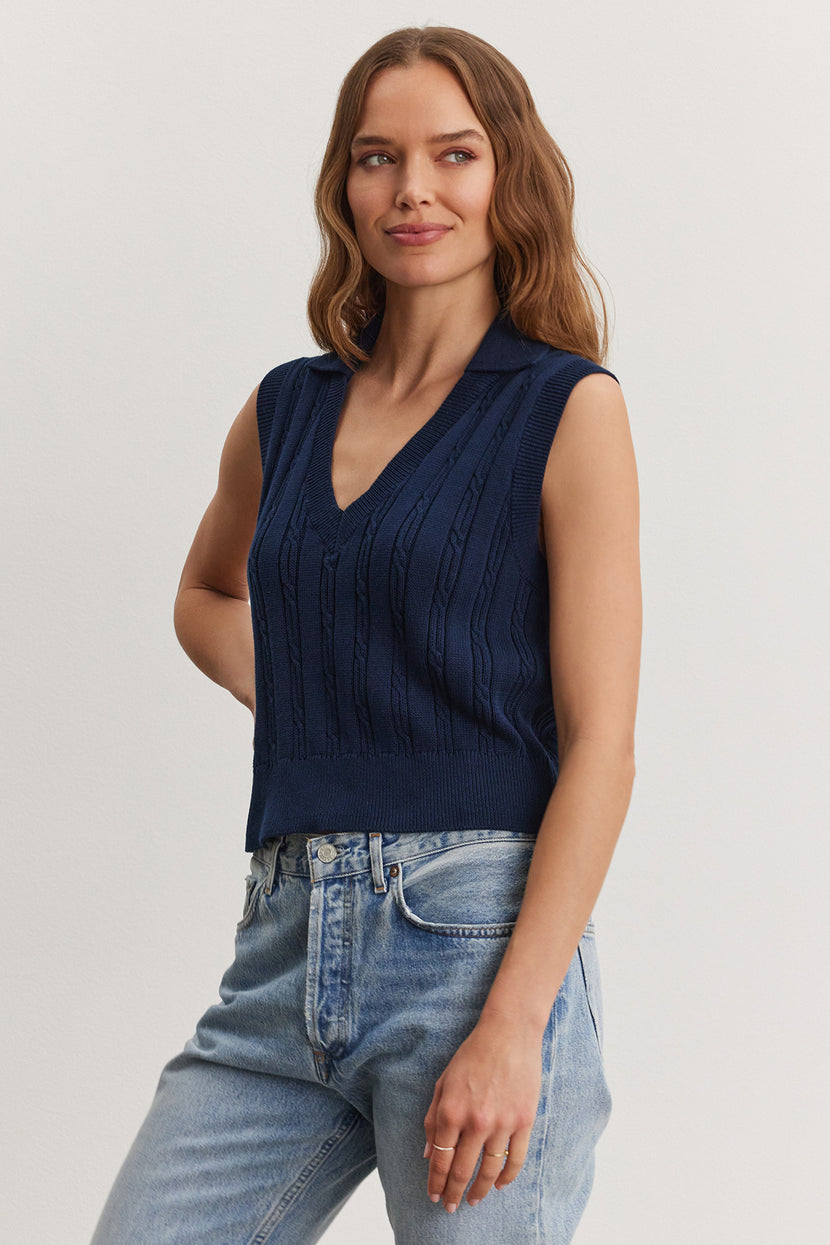 A woman in a Velvet by Graham & Spencer Wendy sweater vest and light blue jeans standing against a plain background, smiling gently.