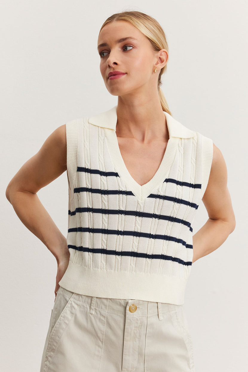 A woman in a cream and navy striped cable knit Wendy sweater vest and cream trousers poses with her hands on her hips against a plain background. (Brand: Velvet by Graham & Spencer)