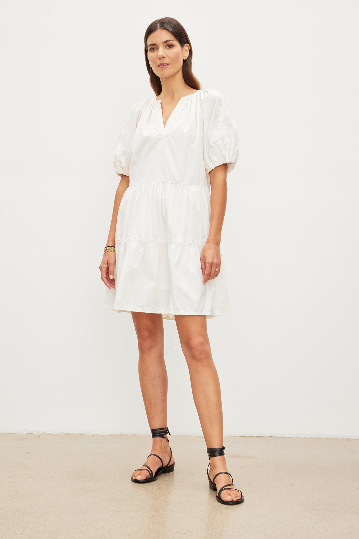   A woman stands in a white room, wearing a white, knee-length cotton dress with puffed sleeves and black sandals, looking directly at the camera. She is wearing the CHRISSY EMBROIDERED BOHO DRESS by Velvet by Graham & Spencer. 