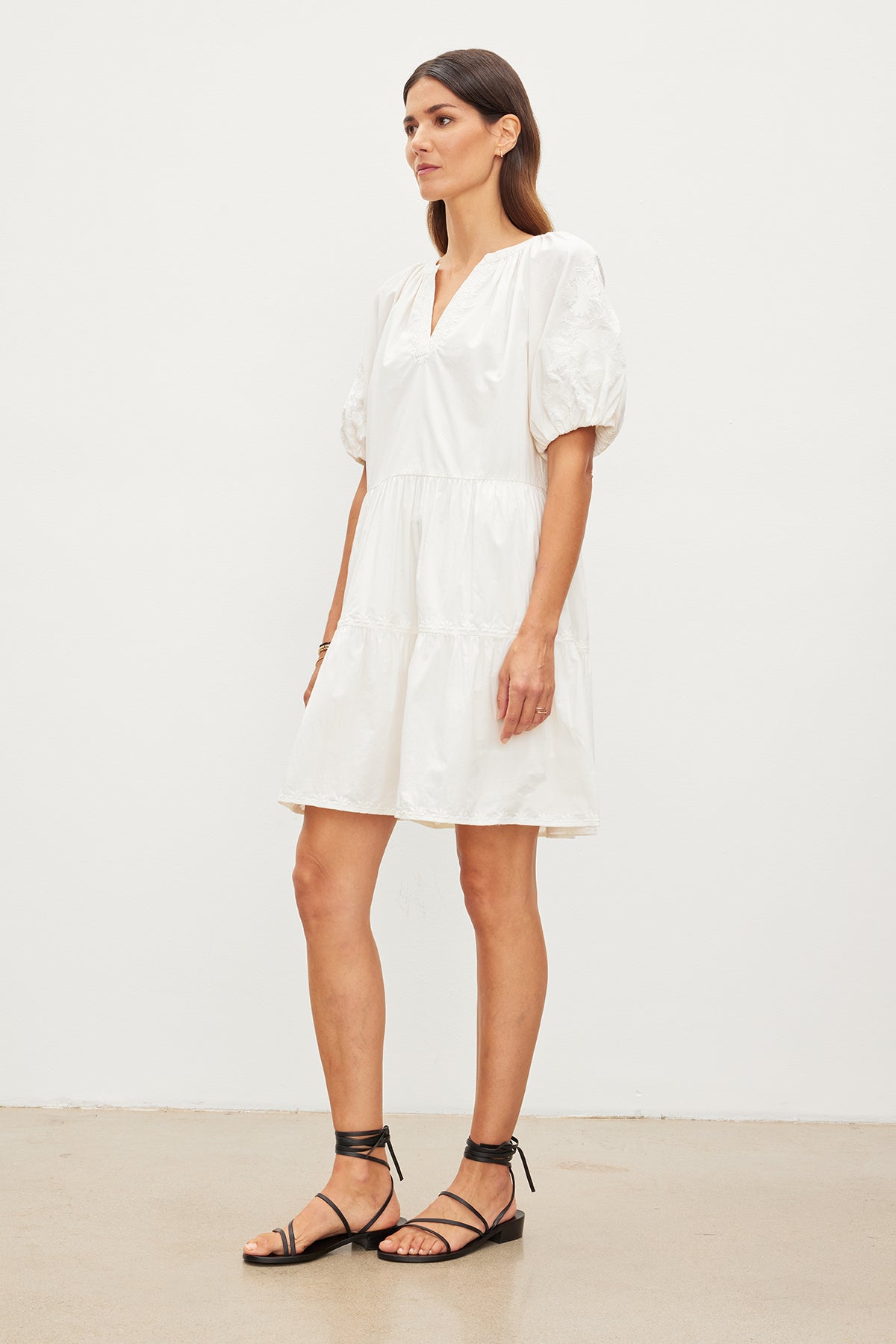 A woman stands in a plain room, wearing a short white cotton CHRISSY EMBROIDERED BOHO DRESS and black strappy sandals from Velvet by Graham & Spencer, looking to the side.-35955557499073
