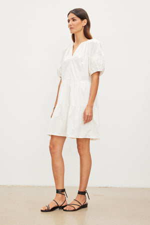 A woman stands in a plain room, wearing a short white cotton CHRISSY EMBROIDERED BOHO DRESS and black strappy sandals from Velvet by Graham & Spencer, looking to the side.