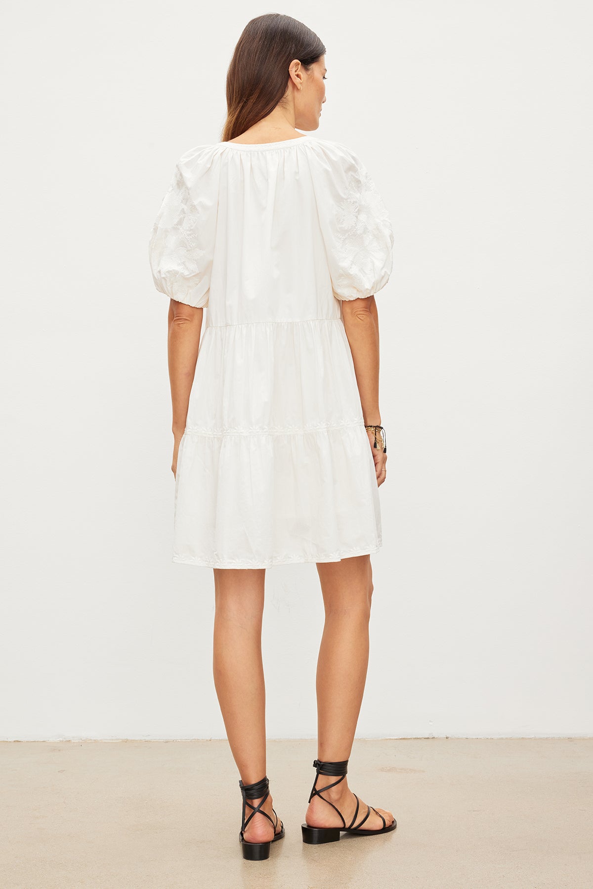   Woman standing with her back to the camera, wearing a white puff-sleeve CHRISSY EMBROIDERED BOHO DRESS by Velvet by Graham & Spencer and black strappy sandals, in a neutral-colored room. 