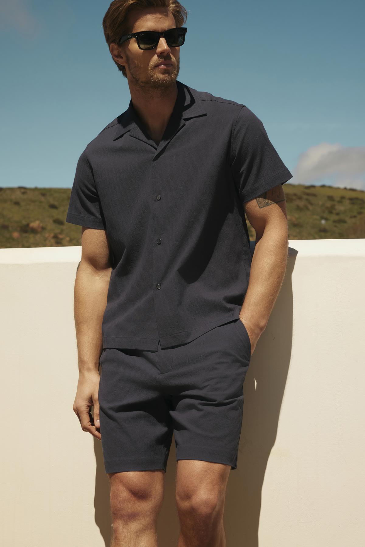 A man in sunglasses wearing a dark shirt and Velvet by Graham & Spencer Damian shorts stands against a white ledge with a clear blue sky in the background.-36753560502465