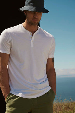 A man wearing a white polo shirt and green Velvet by Graham & Spencer cotton twill FIELDER SHORTS with a blue bucket hat, standing outdoors, with a clear blue sky and ocean in the background.