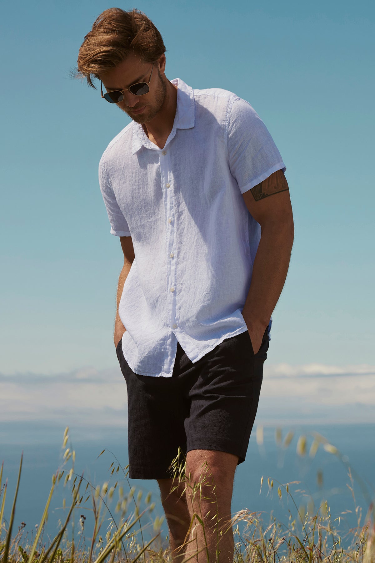 A man in sunglasses, wearing a light blue shirt and Velvet by Graham & Spencer's seersucker cotton Damian shorts, stands in a field overlooking the sea on a sunny day.-36909331742913