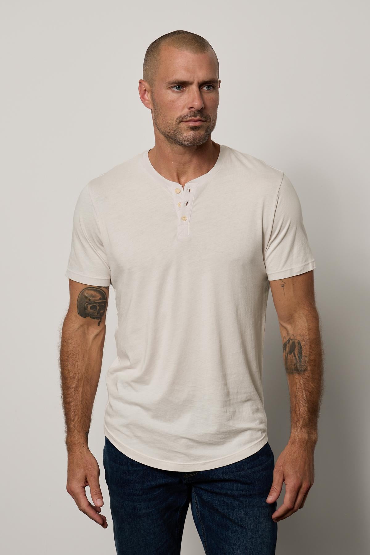 A bald man with tattoos on both arms, wearing a white Velvet by Graham & Spencer Fulton Henley with a curved hemline, standing against a neutral background.-36890721943745