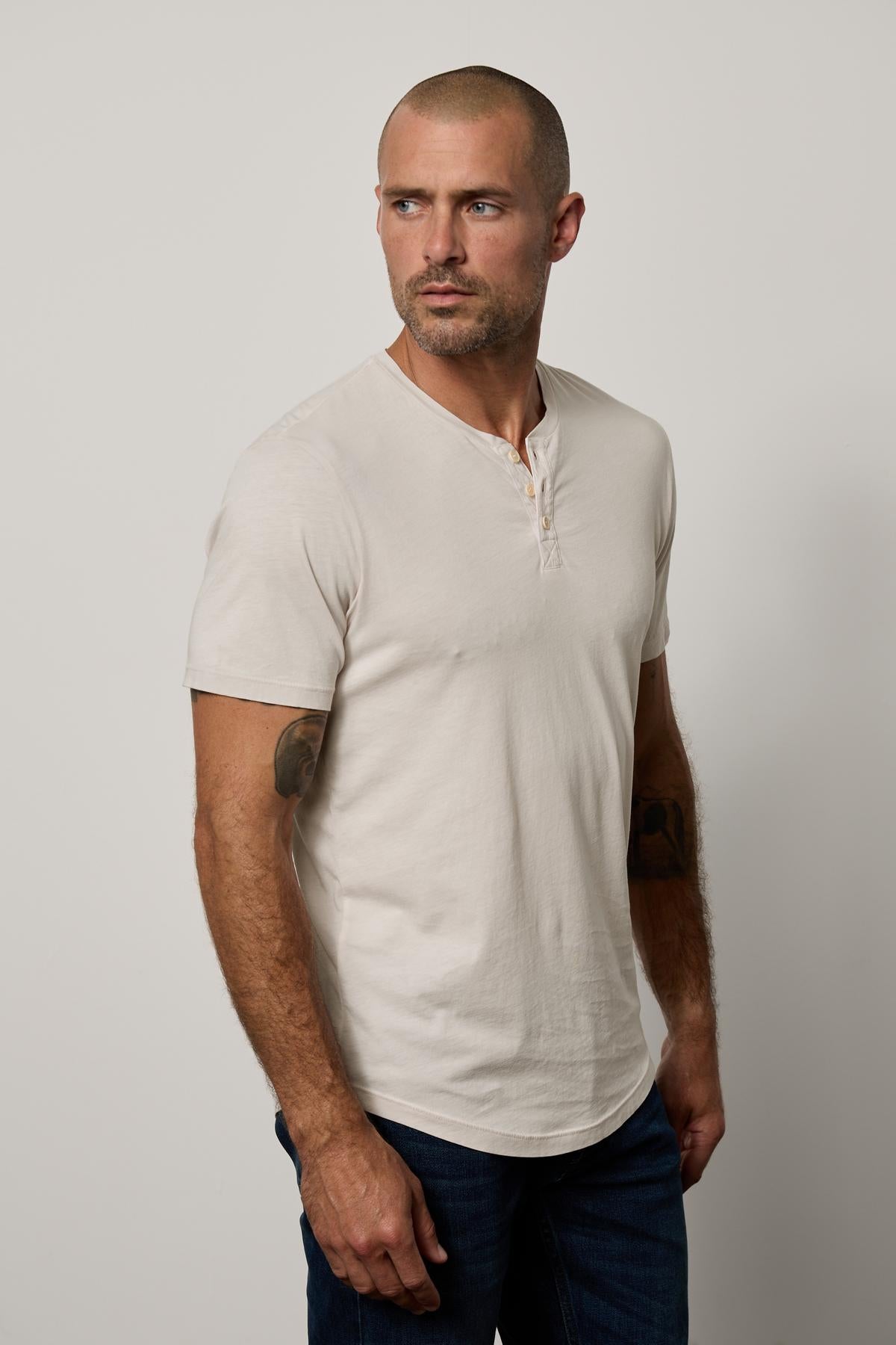   A man with a short haircut and tattoos on his arms stands facing left in a Velvet by Graham & Spencer Fulton Henley tee against a white background. 