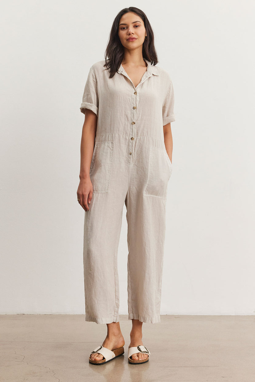 A woman models a casual beige Velvet by Graham & Spencer DELILAH LINEN JUMPSUIT with short sleeves and front buttons, paired with white sandals, standing in a simple studio setting.